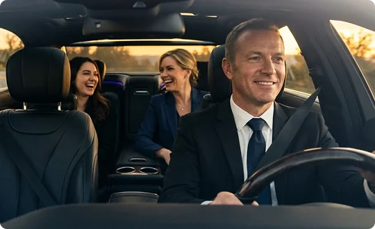 A smiling chauffeur in a professional black suit and tie drives a luxury vehicle with black leather seats. In the back seat, two well-dressed female passengers are laughing and enjoying their conversation during the ride.