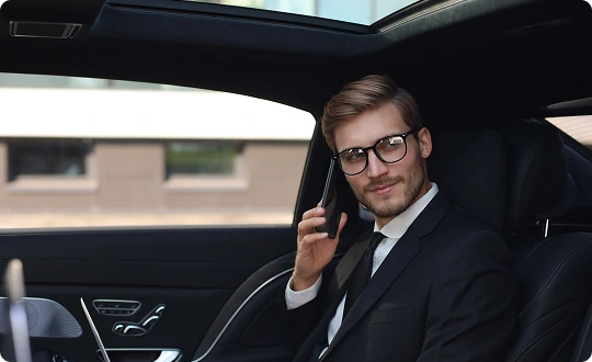 Stylish businessman in glasses making a phone call inside a chauffeur-driven car with a panoramic sunroof and leather interior