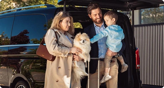 A smiling family with a small dog and a young child standing beside a black van with its rear door open.