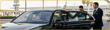 Two professional chauffeurs, both dressed in suits, attend to a black luxury sedan at an airport or terminal. One chauffeur is holding the rear door open, while the other stands ready, with palm trees and modern buildings visible in the bright background.